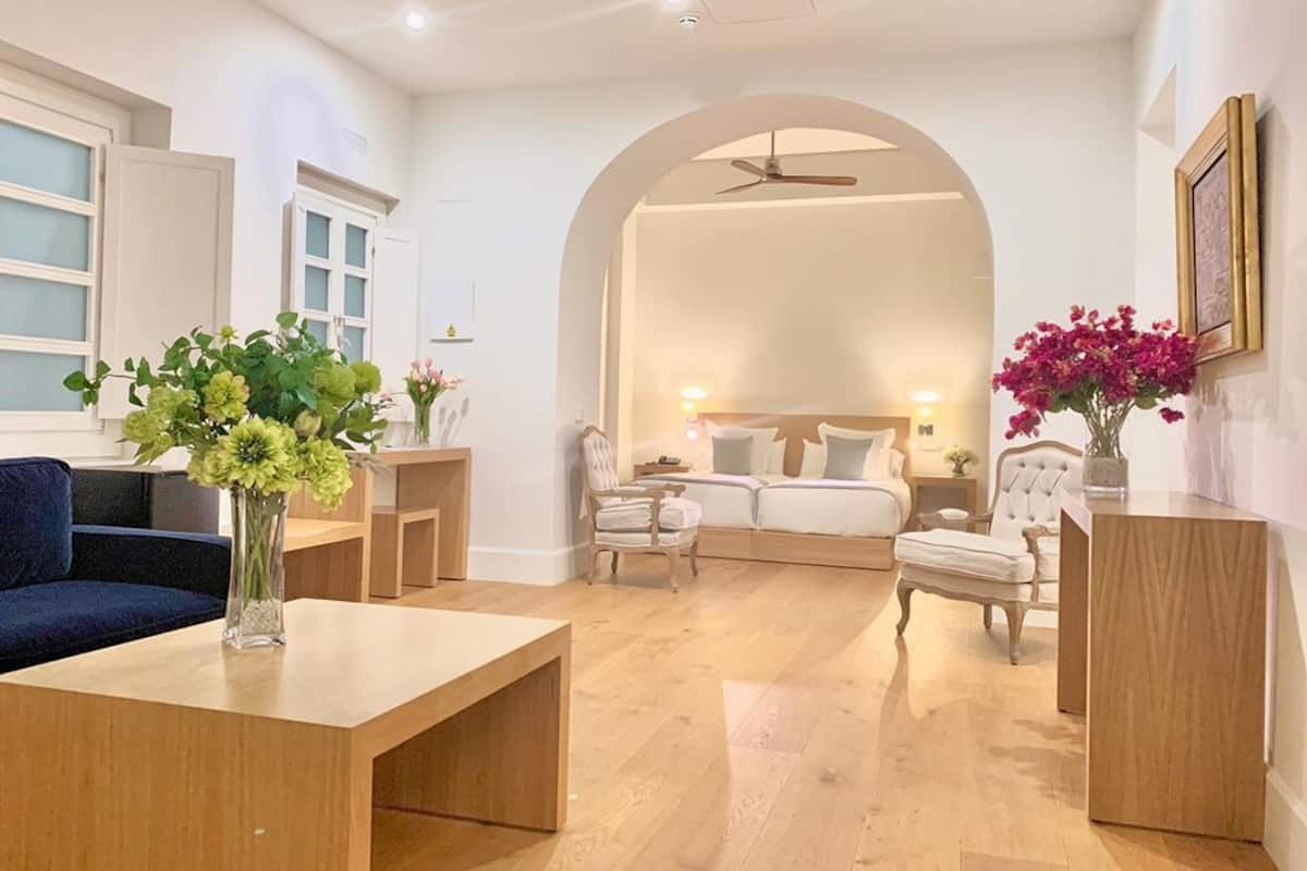 A modern living room with a sofa, a coffee table, and a dining table. There is a bed with a headboard in the background.