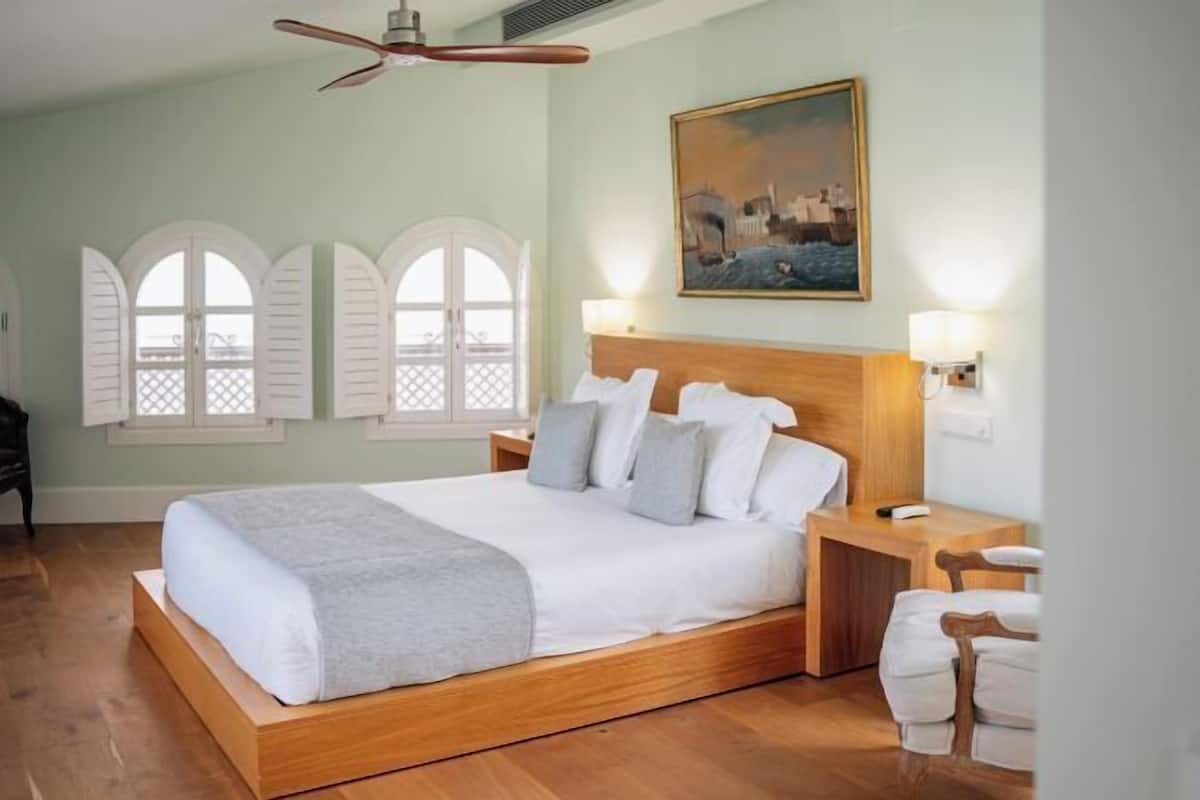 A bedroom with a wooden bed, a nightstand, a chair, a ceiling fan, and a painting on the wall.