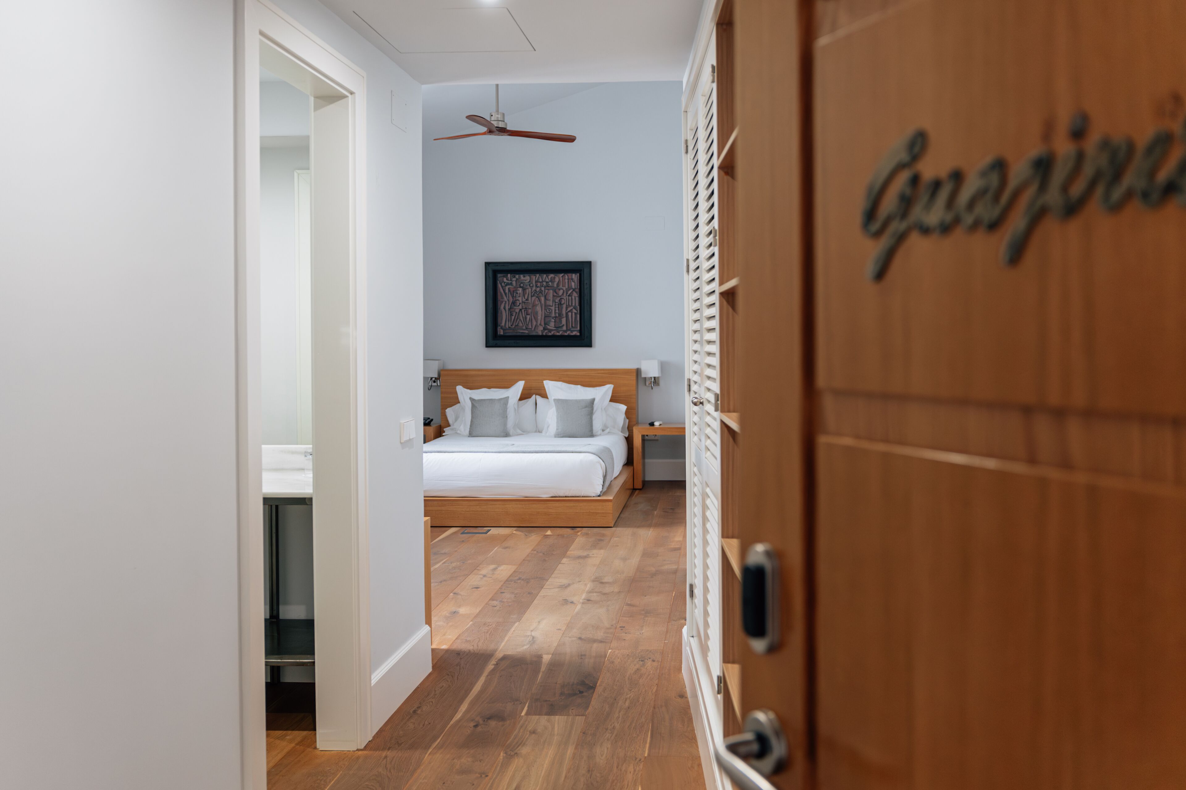 Une chambre avec un plancher en bois, un lit avec une literie blanche, un ventilateur de plafond et un tableau encadré au mur.