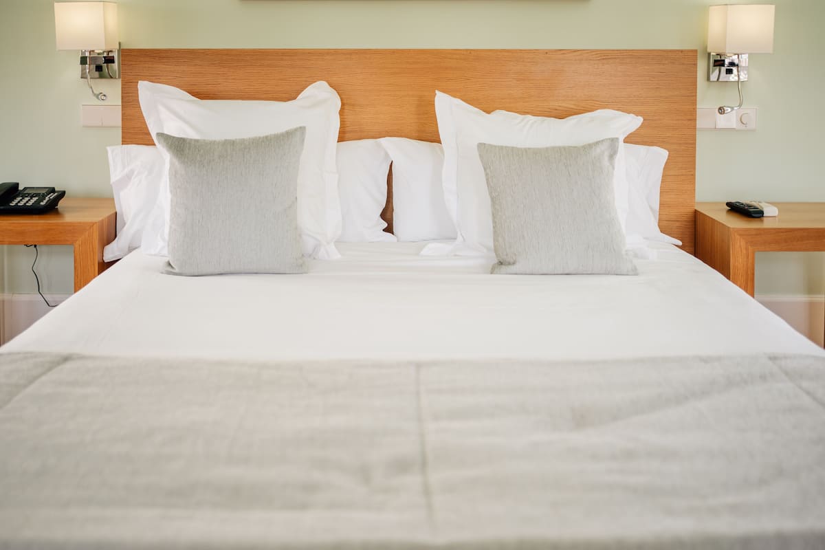 A neatly made bed with white linens and two pillows, a wooden headboard, bedside tables with lamps, and a phone on one table.