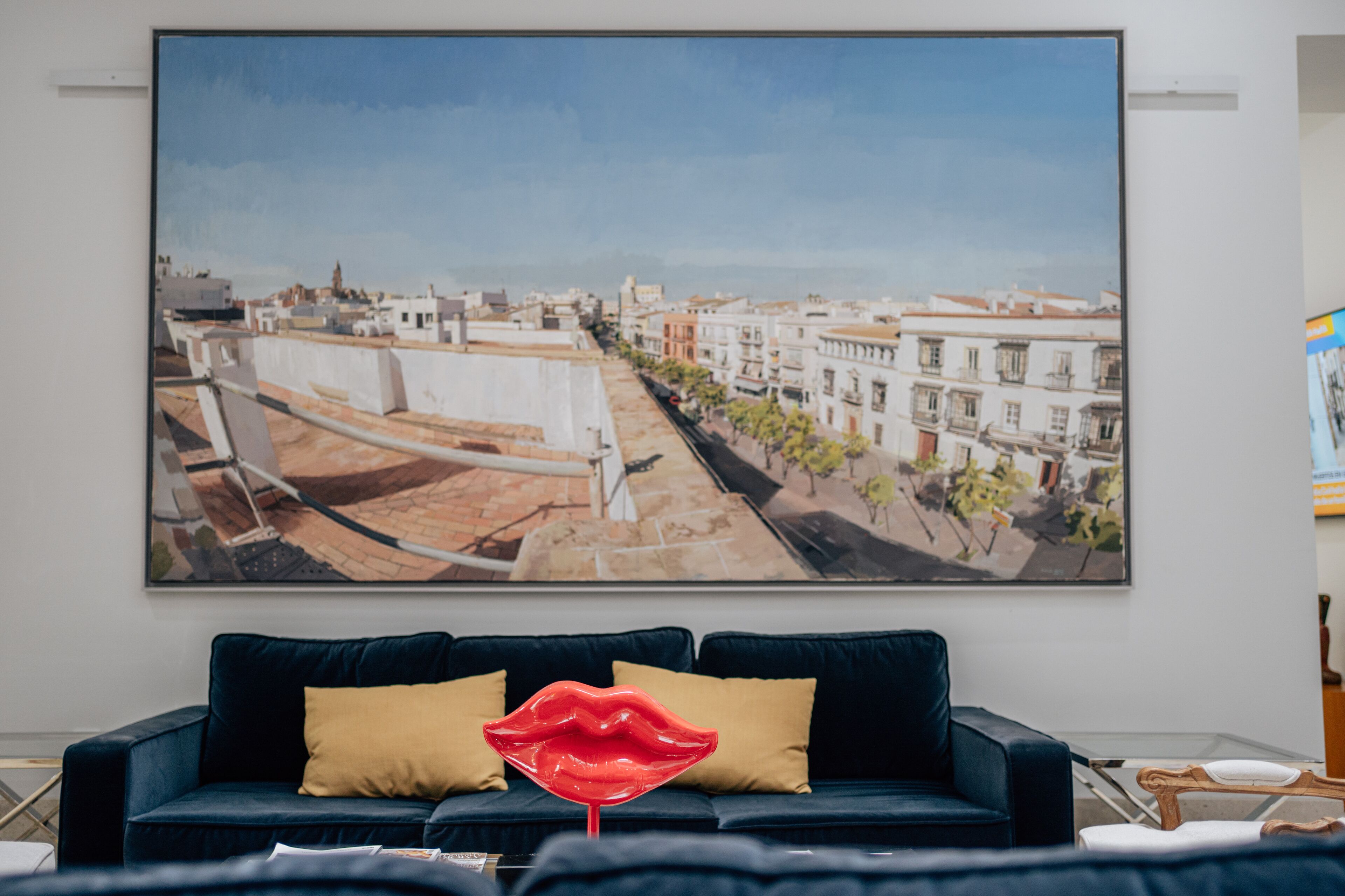 A living room with a large cityscape painting, a dark blue sofa with yellow cushions, and a red lips decoration.