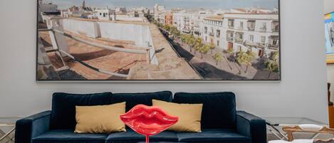 A living room with a large cityscape painting, a dark blue sofa with yellow cushions, and a red lips decoration.