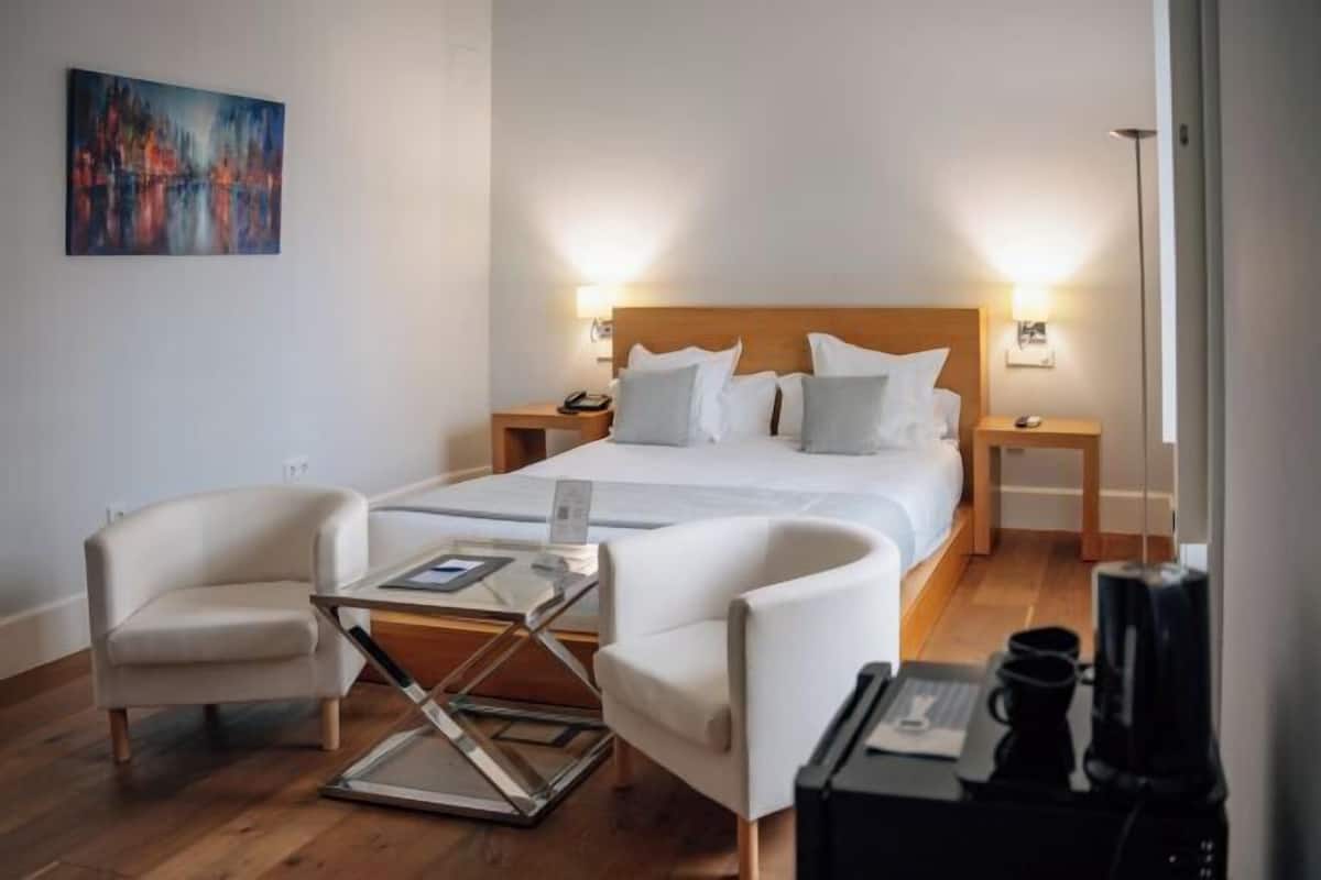 A hotel room with a bed, two bedside tables, a desk with a chair, a coffee machine, and a painting on the wall.