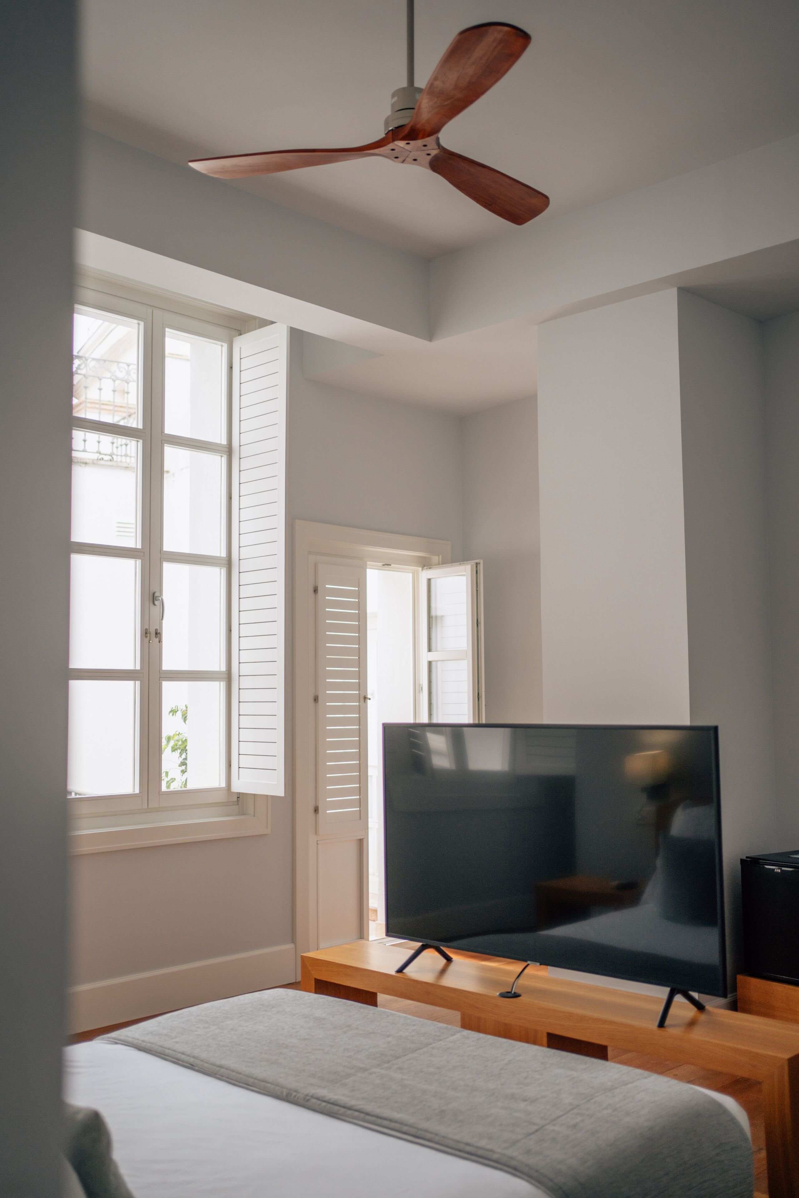 Un dormitorio con cama, televisor sobre un mueble de madera, ventilador de techo y ventana con persianas.