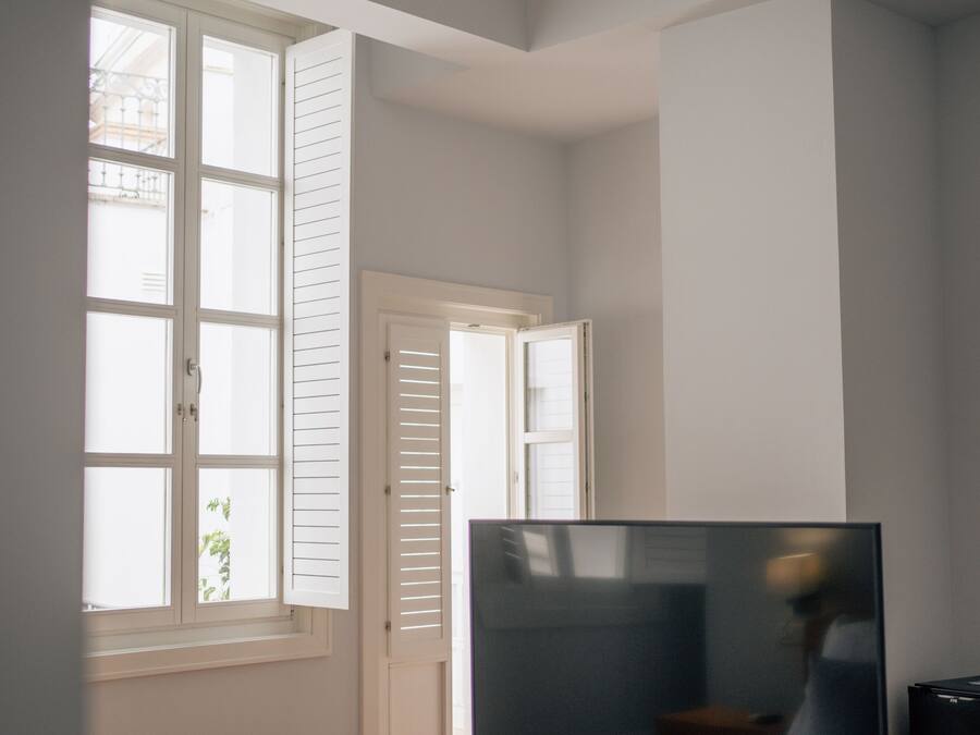 Un dormitorio con cama, televisor sobre un mueble de madera, ventilador de techo y ventana con persianas.