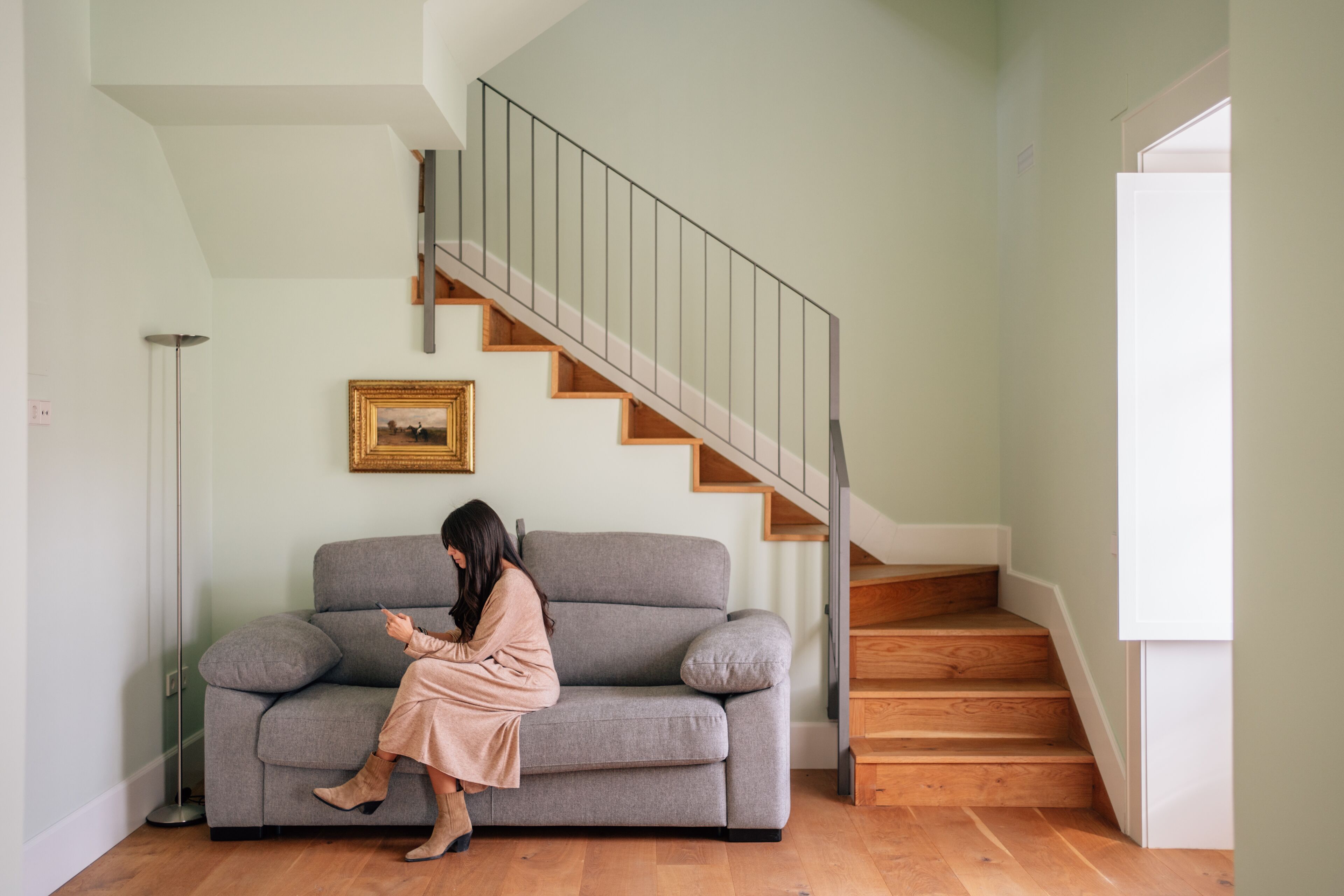 Una mujer sentada en un sofá gris en una habitación con una escalera de madera y un cuadro en la pared.