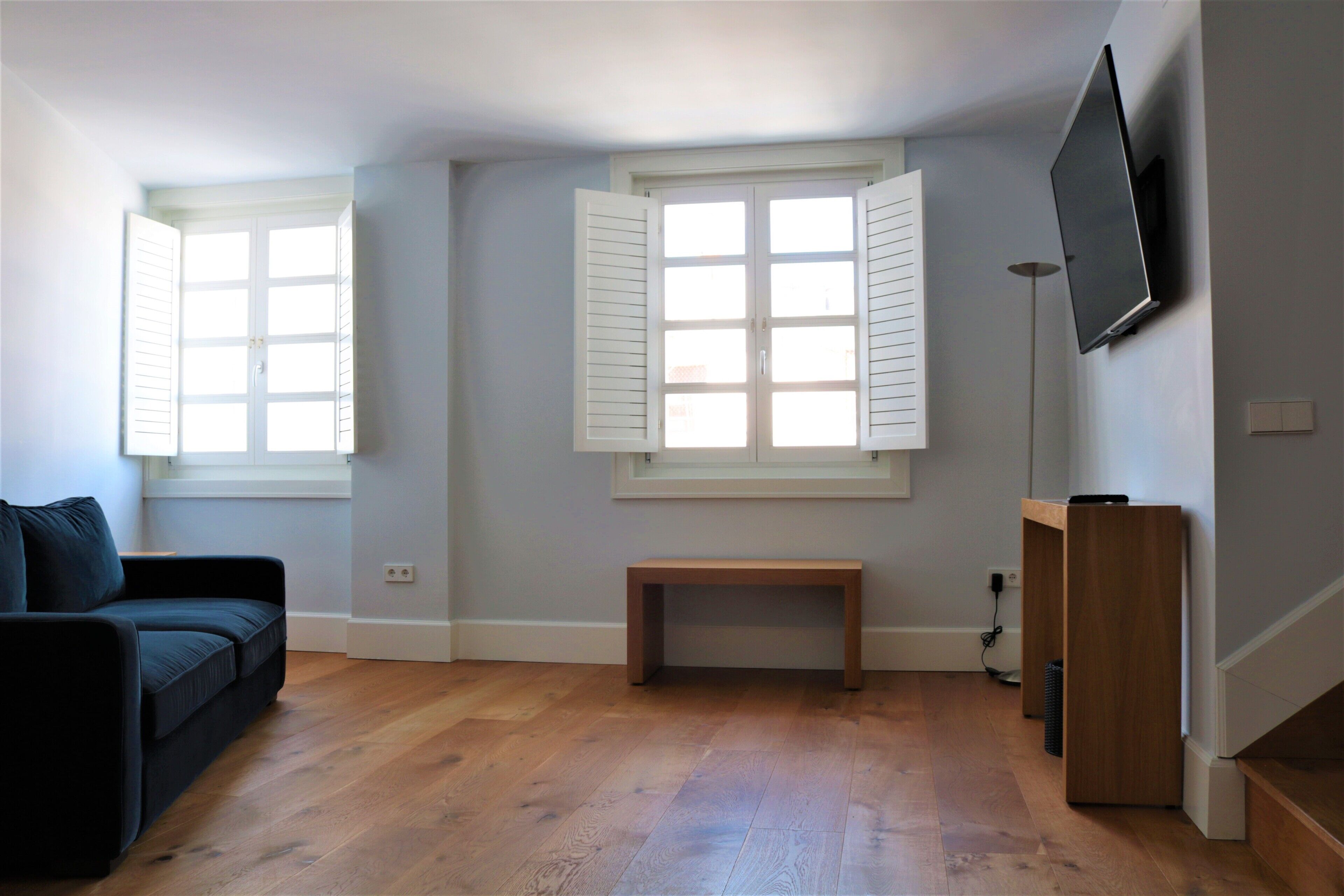 A living room with a dark-colored sofa, a wooden bench, a flat-screen TV mounted on the wall, and a window with white shutters.