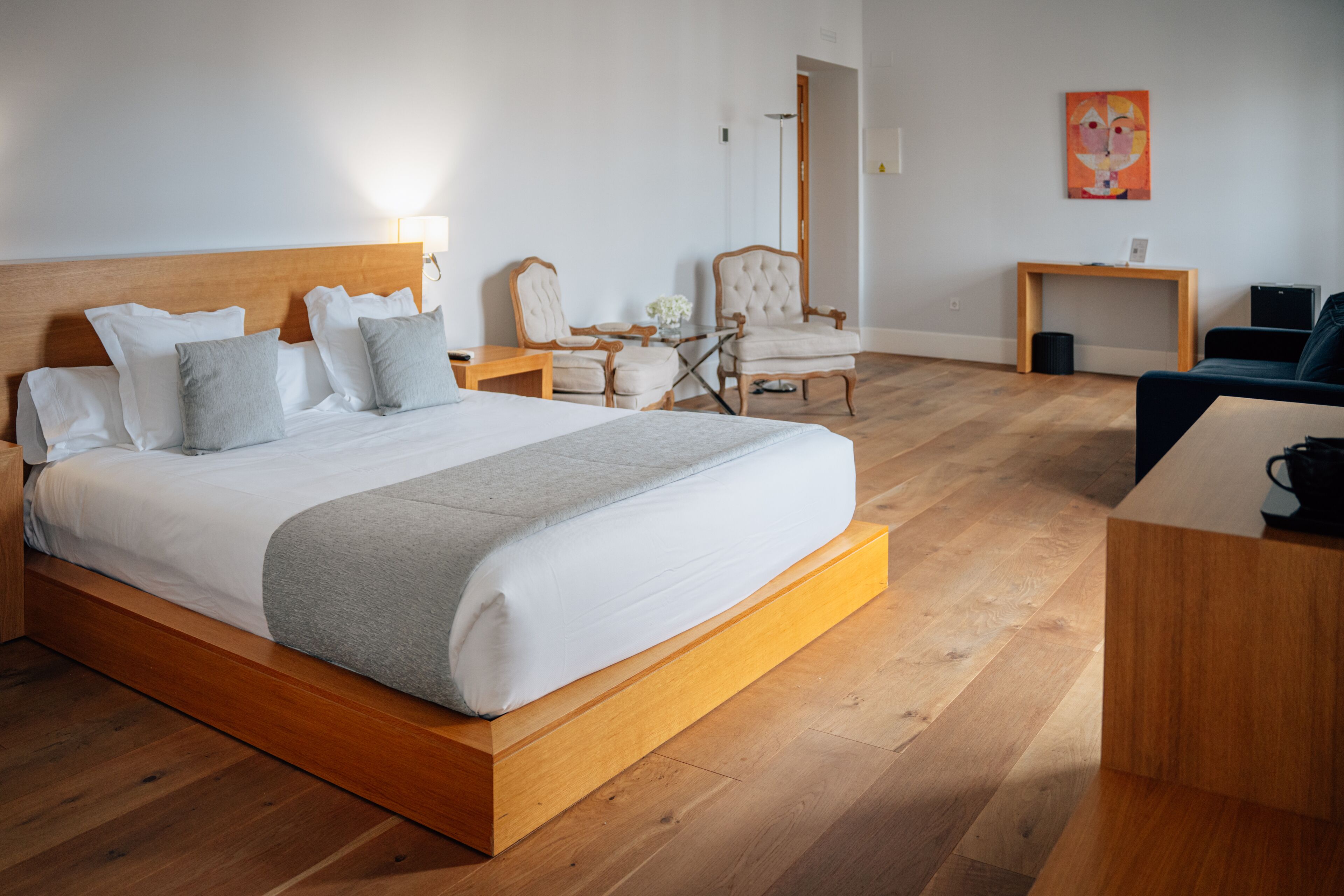 A modern hotel room with a large bed, a wooden headboard, a desk, a chair, and a small table.