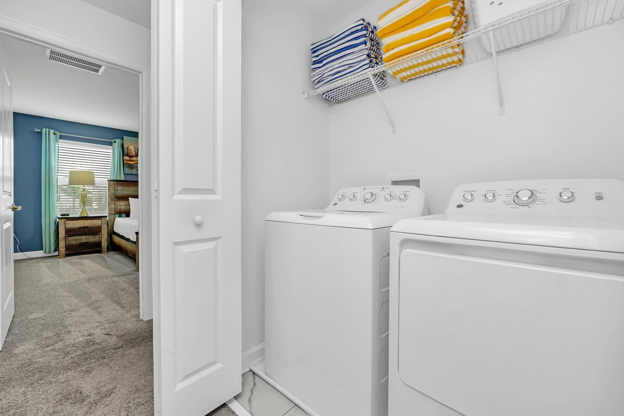 Una sala de lavandería con dos lavadoras blancas, un tendedero con ropa doblada y una puerta que da a un dormitorio con una cama y una mesita de noche.