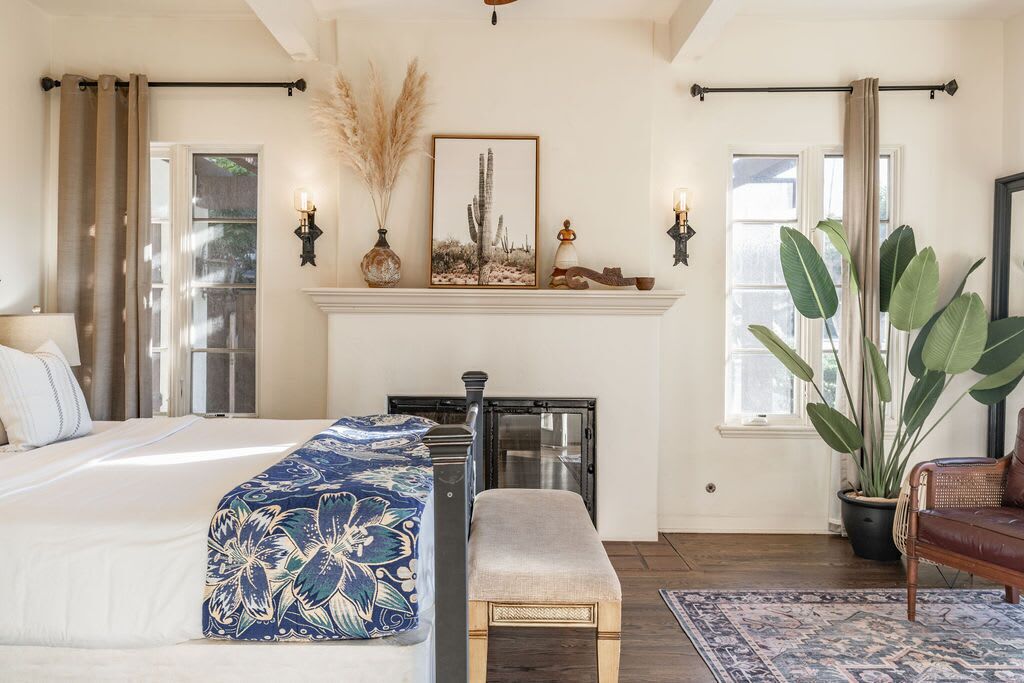 A bedroom with a bed, bench, large plant, fireplace, and windows.