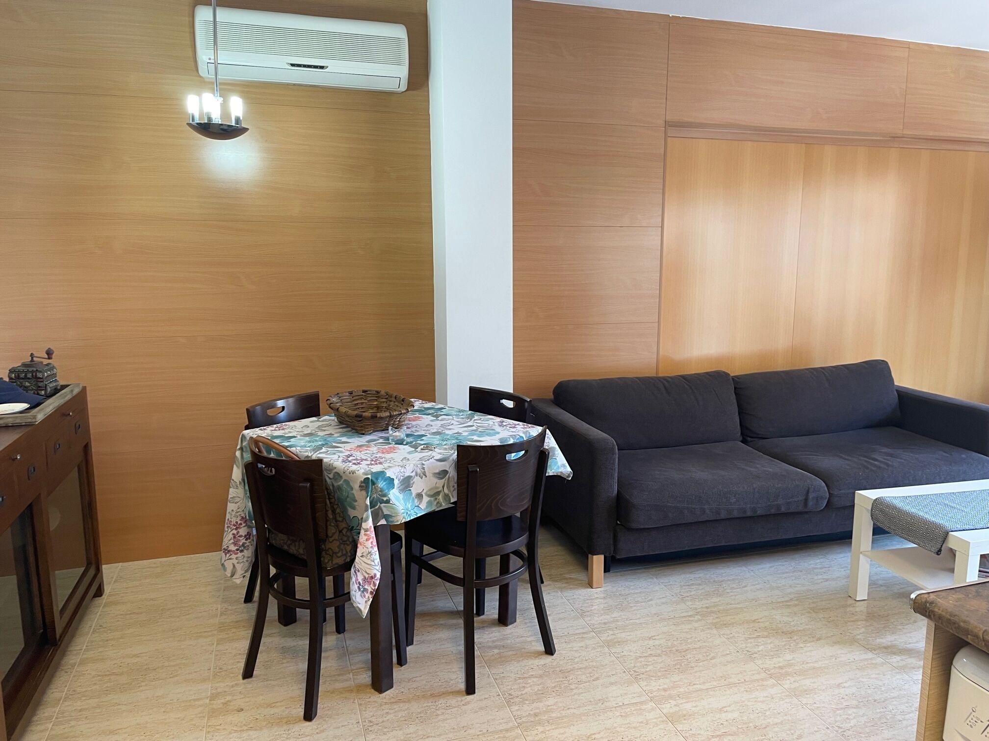 Un comedor con una mesa dispuesta para cuatro personas, un sofá de color oscuro y una mesita con una cesta.