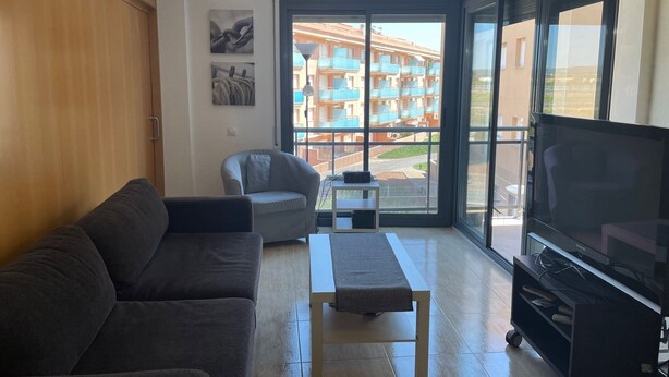 Een woonkamer met een bank, een stoel, een klein tafeltje en een televisie. Er is een raam met uitzicht op een gebouw met balkons.