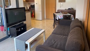 Een woonkamer met een bank, een televisie en een kleine tafel. Achtergrond is een keuken met een koelkast en een eettafel.