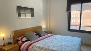 Een net opgemaakt bed met een geborduurd sprei, twee kussens en een bedlampje op een houten nachtkastje. Daarboven hangt een ingelijst fotoprint aan de muur.