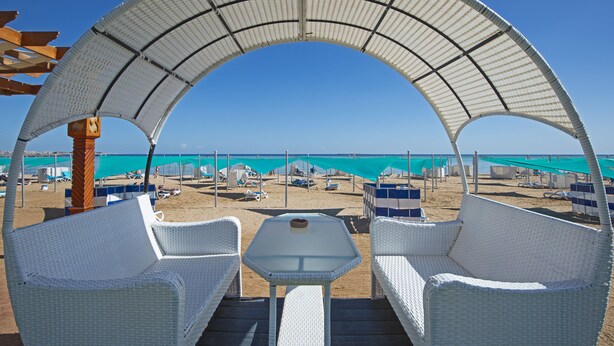 Un espace aménagé avec des chaises en osier blanc et une table, situé en bord de plage et offrant une vue sur la plage et les eaux turquoise.