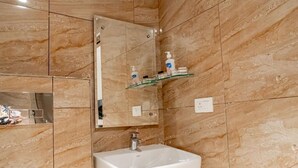 Un baño con pared de mármol, un lavamanos blanco, un espejo y un estante de vidrio con artículos de aseo.