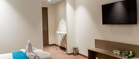 A hotel room with a bed, a TV mounted on the wall, a small table, and a bench with a tray of items.