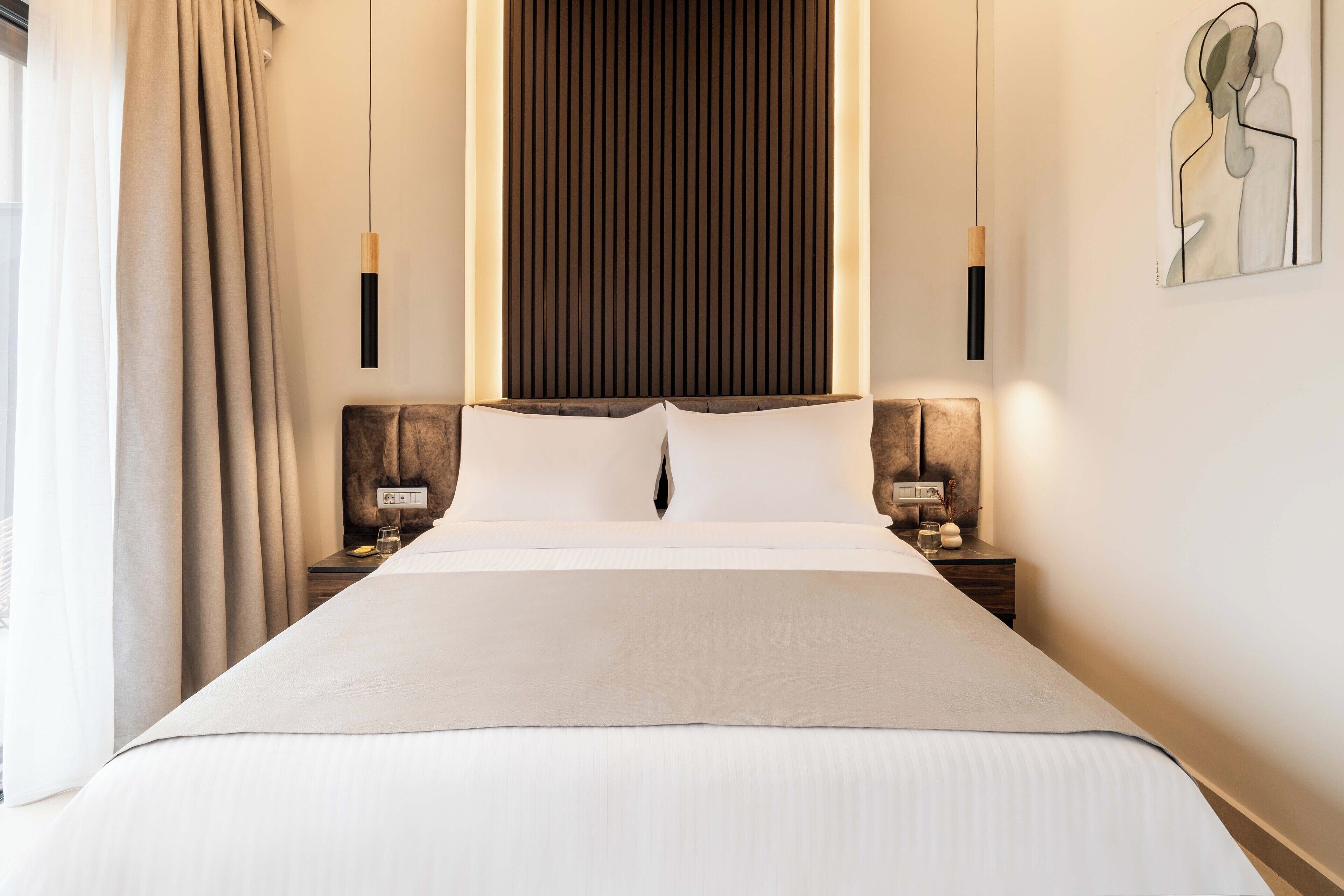 A neatly made bed in a hotel room with a large window, two bedside tables, and a wall-mounted artwork.