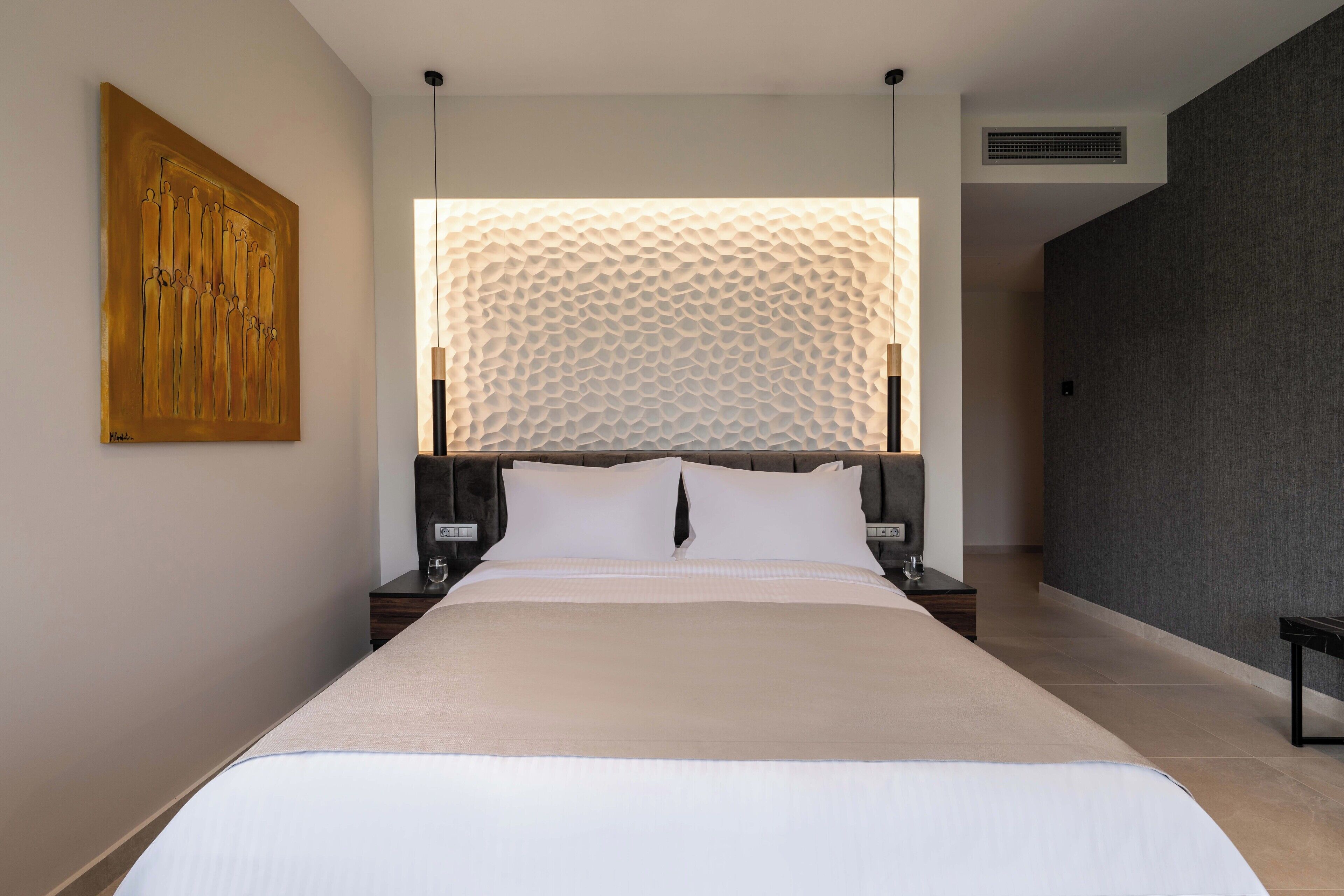 Une chambre moderne avec un grand lit, une tête de lit au motif texturé, une œuvre d’art accrochée au mur et une table de chevet avec une lampe.