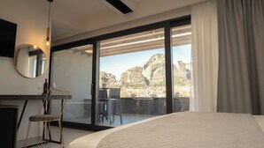 A modern hotel room with a large bed, a desk with a chair, a large mirror, and a sliding glass door leading to a balcony with a view of rocky mountains.
