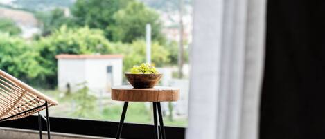 Une vue depuis un balcon, avec une chaise en osier et un tabouret en bois sur lequel est posé un vase rempli de fleurs jaunes.