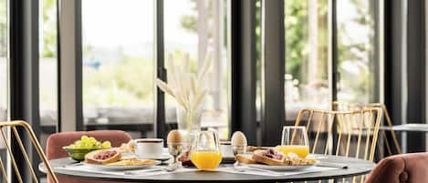 A breakfast table set with eggs, toast, fruit, and orange juice in a modern dining area with large windows and metal chairs.