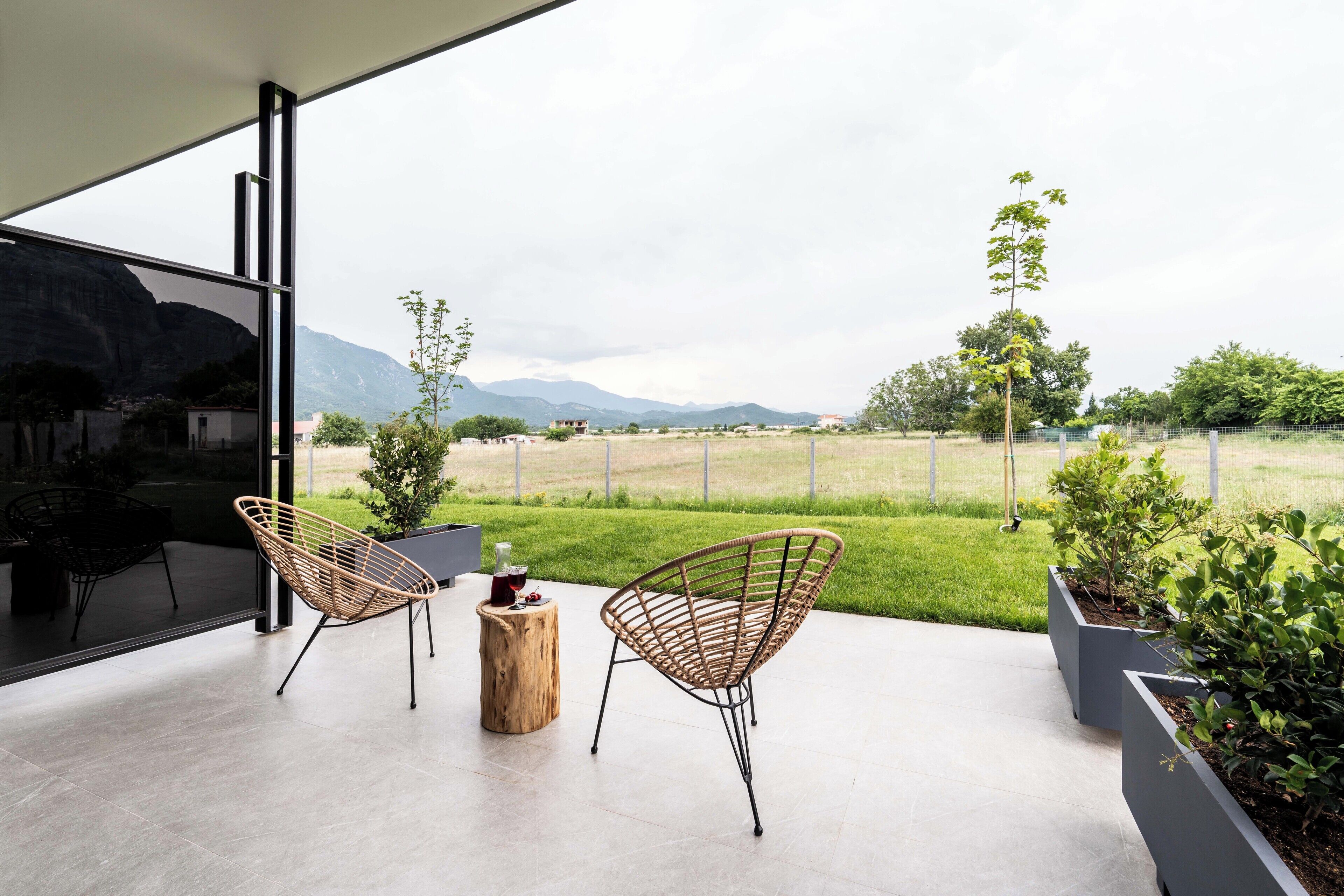 A modern outdoor patio with two wicker chairs, a wooden stool, and a glass table. A large window offers a view of a green field and distant mountains.