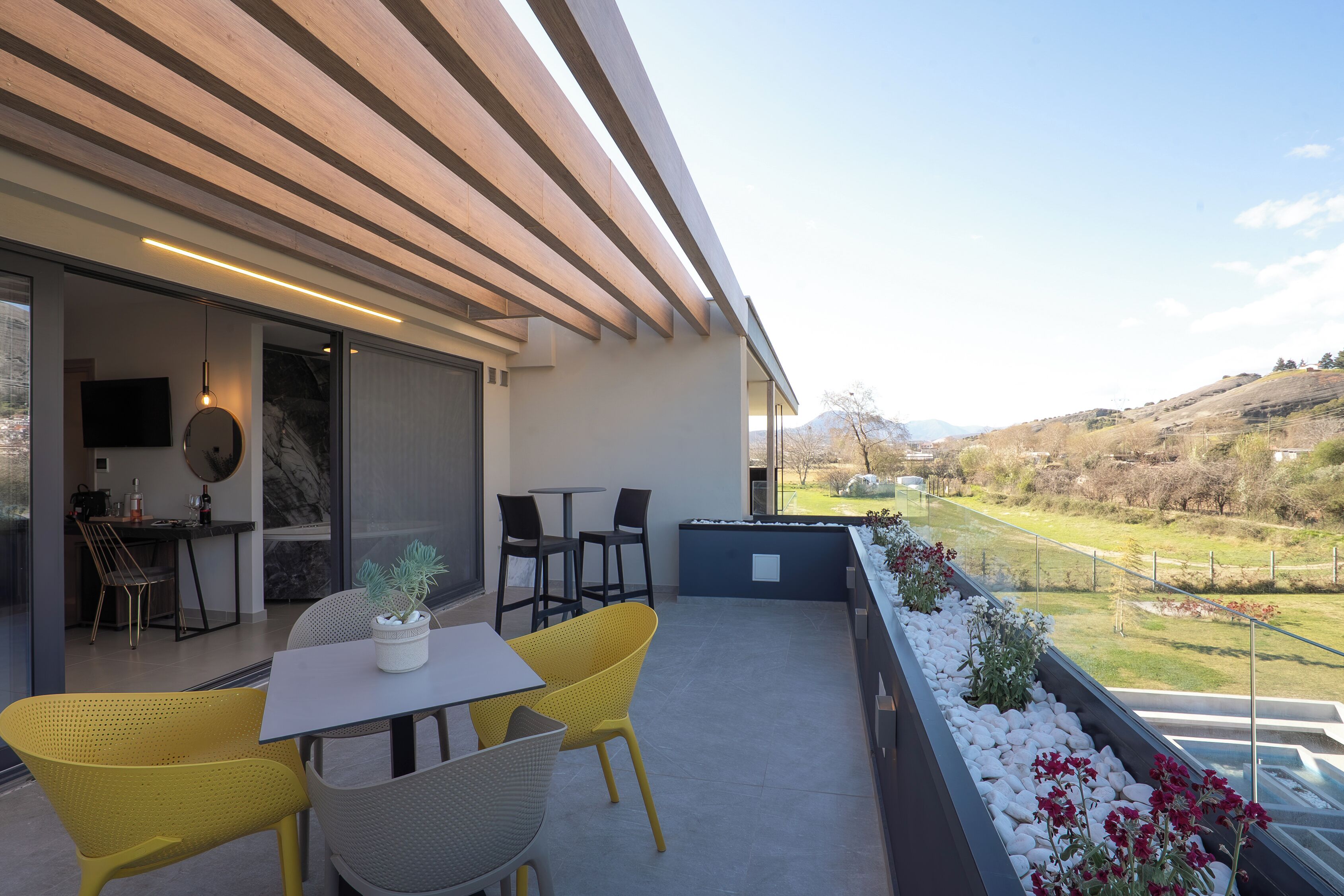 A modern outdoor patio with a table and chairs, a potted plant, and a view of a green landscape.