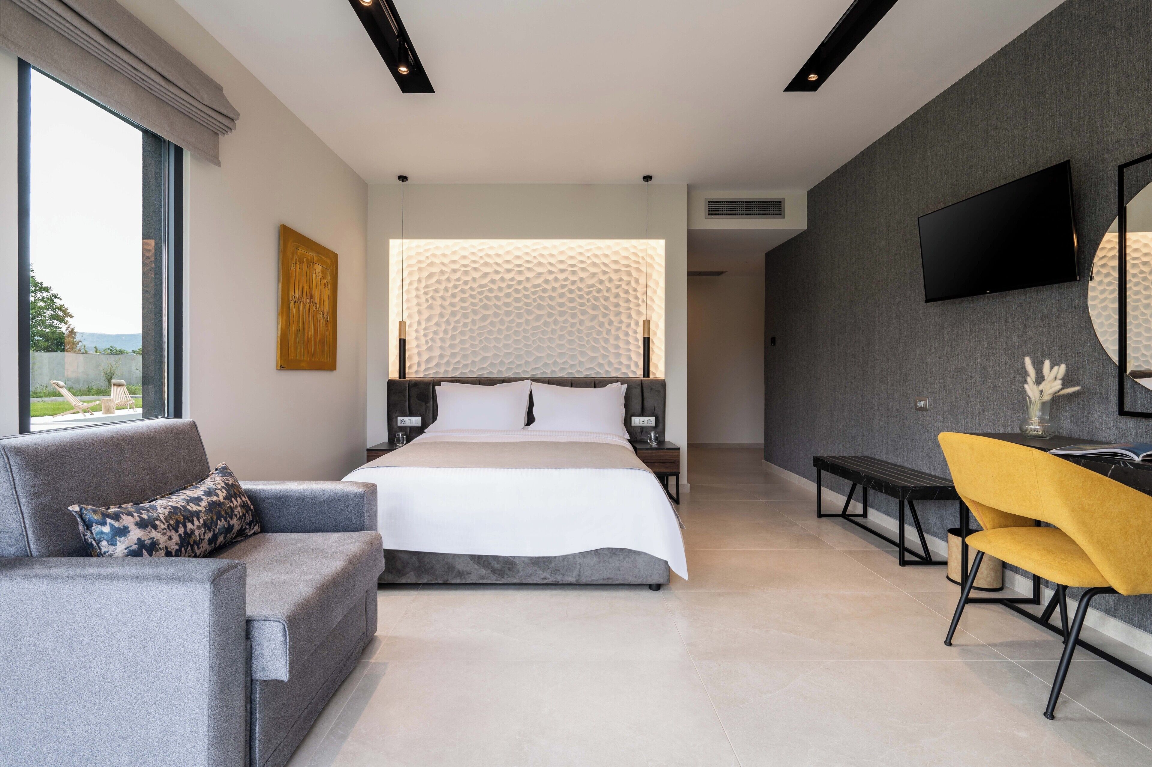 Un dormitorio moderno con una cama grande, un sofá gris, un sillón amarillo y un televisor montado en la pared.