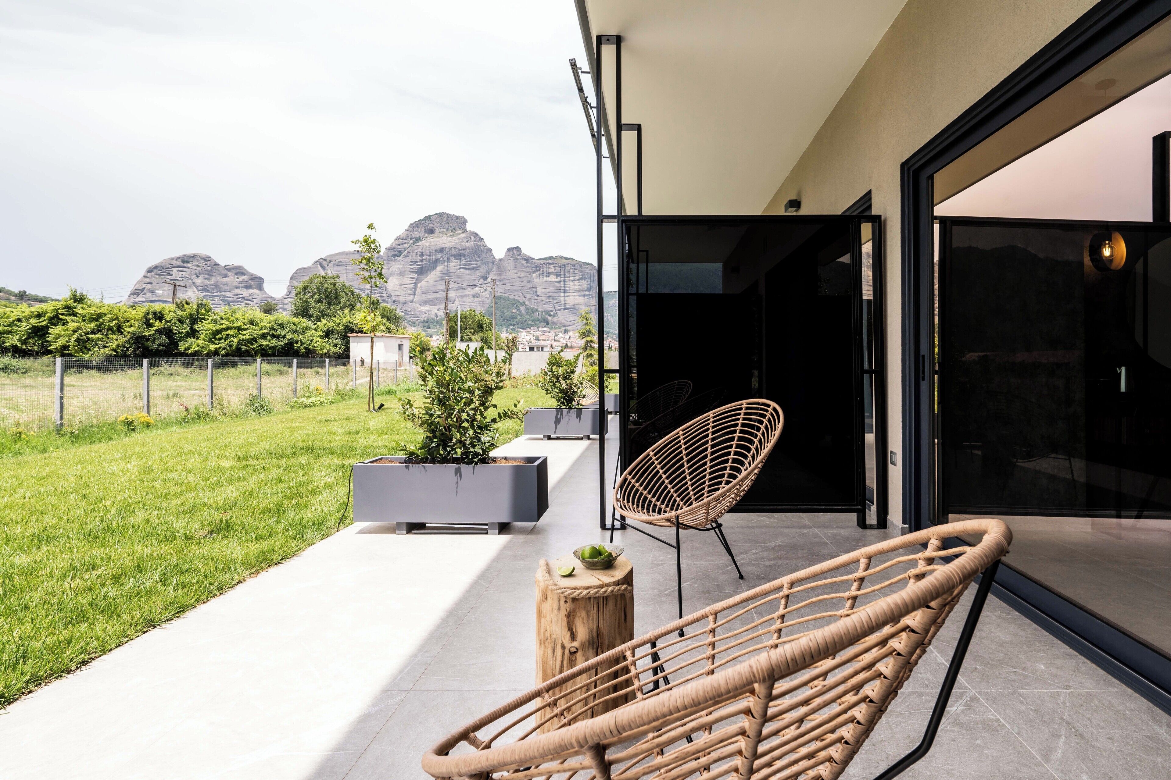 Un patio al aire libre con mobiliario de mimbre, la entrada de un edificio moderno y vistas a montañas distantes.