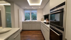 A modern kitchen with white cabinets, a built-in oven, and a window with a view of greenery. - Two Bedroom Flat in Belgravia (London)