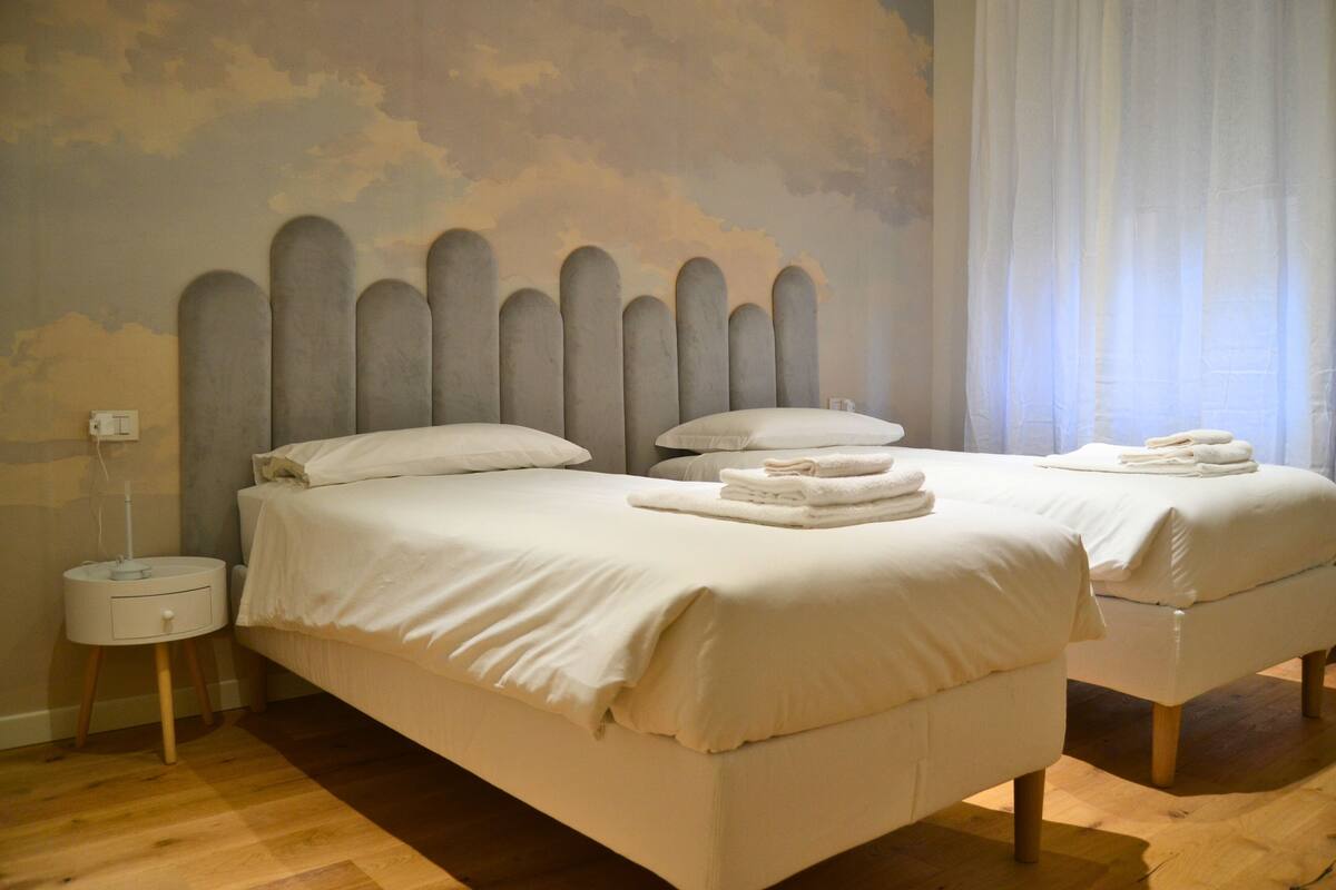 Une chambre avec deux lits, une table de chevet et un mur décoré d’une fresque représentant des nuages.