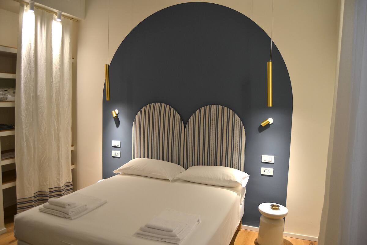 Un lit bien fait, avec du linge de lit blanc, une tête de lit sombre à rayures verticales et une petite table de chevet surmontée d’une lampe blanche.