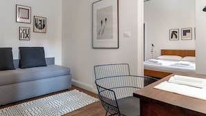 Ein moderner, gut beleuchteter Raum mit grauem Sofa, einem schwarzen Drahtgestellstuhl, einem Holztisch mit einem Buch und einem Bett mit weißen Leinen.