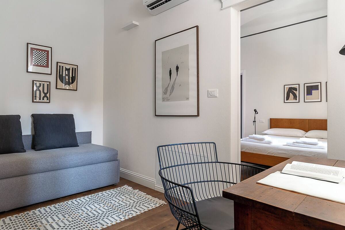 Une pièce moderne et bien éclairée, avec un canapé gris, une chaise en fil de fer noir, un bureau en bois sur lequel se trouve un livre, et un lit recouvert de draps blancs.