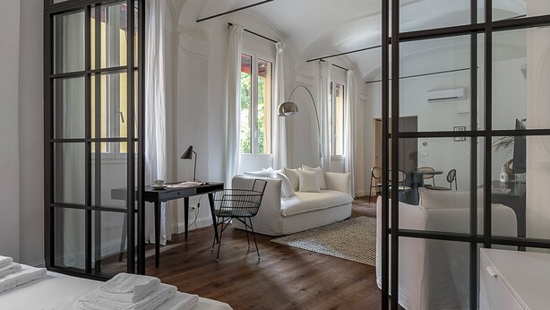 Ein modernes Hotelzimmer mit einem großen Fenster, einem Schreibtisch, einem weißen Sofa und einem Bett mit ordentlich gefalteten weißen Handtüchern.