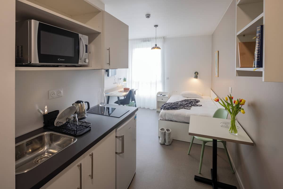 Une chambre d’hôtel compacte comprenant un coin cuisine, un lit, une petite table avec un vase de fleurs et un bureau avec une chaise.
