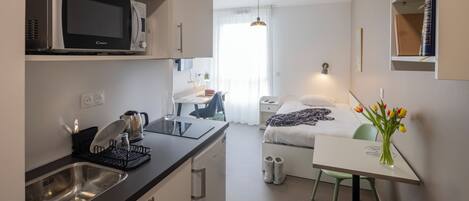 Habitación de hotel compacta con kitchenette, una cama con manta, una mesita de noche, una silla y un pequeño escritorio con lámpara.