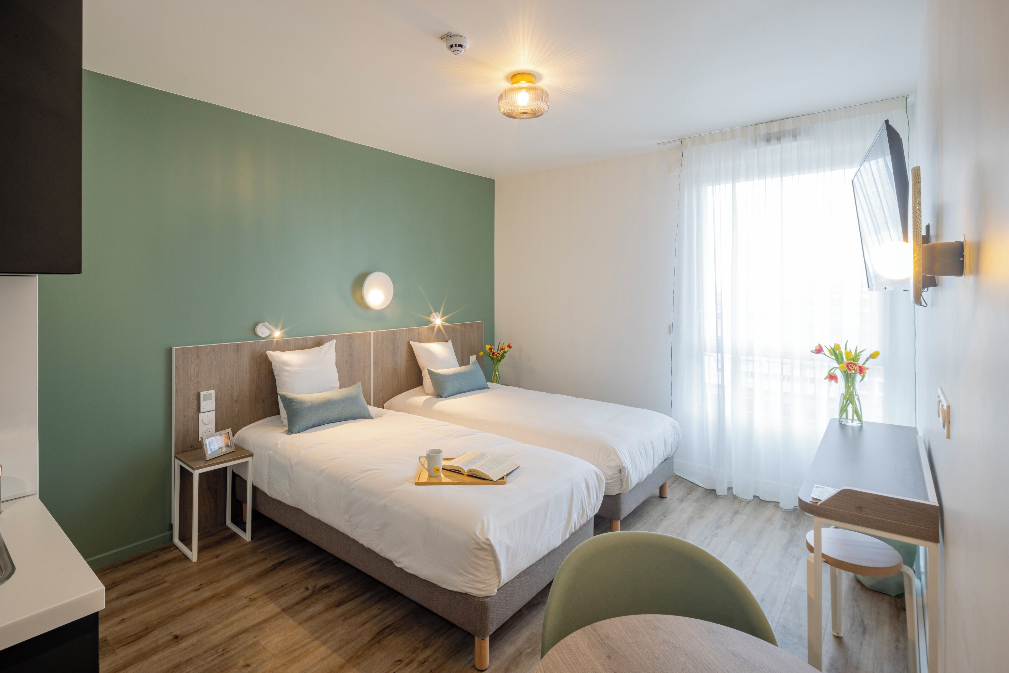 Quarto de hotel com duas camas, uma escrivaninha, uma cadeira, uma TV e uma janela com cortinas.