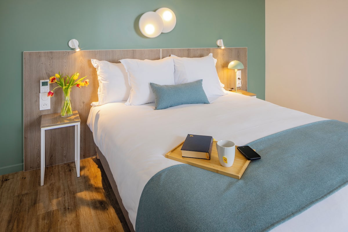 Un lit bien fait, avec du linge de lit blanc, un plaid bleu et une table de chevet sur laquelle se trouvent un livre, une tasse et un téléphone intelligent.