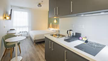 Habitación de hotel compacta con kitchenette, una cama, un pequeño comedor y una ventana con cortinas.