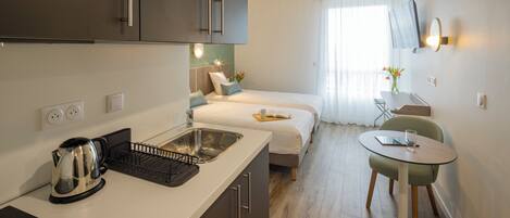 A modern hotel room with a kitchenette, a bed, a small dining area, and a TV mounted on the wall.