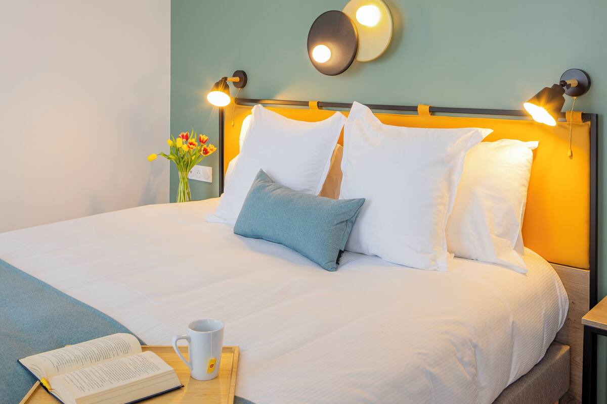Un lit bien fait, avec une literie blanche et bleue, une table de chevet sur laquelle se trouvent un livre et une tasse, et des lampes fixées au mur.