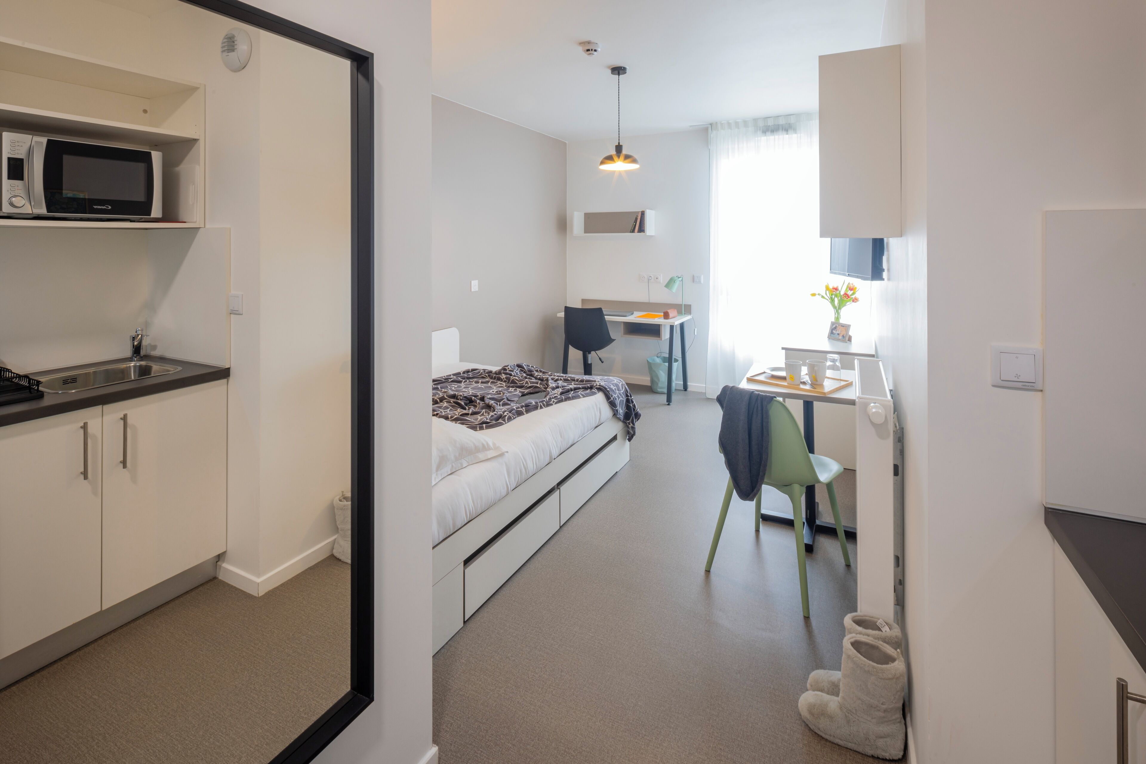 A compact hotel room with a kitchenette, a bed with a patterned blanket, a desk with a chair, and a small table with a lamp.