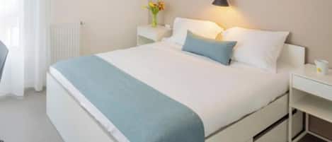 A neatly made bed with a white and blue bedspread, a bedside table with a lamp, and a vase with flowers on the nightstand.