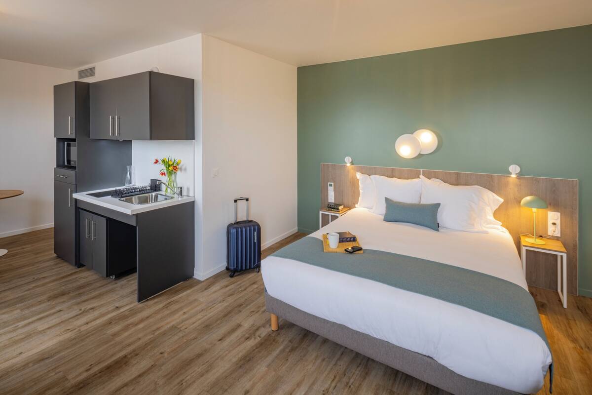 Une chambre d’hôtel moderne dotée d’une kitchenette, d’un grand lit avec une tête de lit en bois et d’un mur d’accent vert.