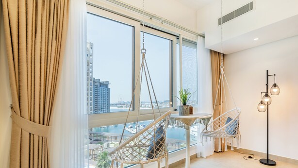 A modern room with large windows, a hanging chair, a small table with a plant, and a floor lamp. - Marina Residences 2 501 (Dubai)
