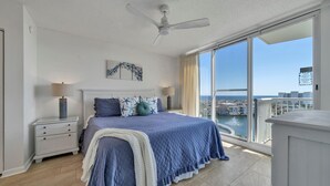 Ein Schlafzimmer mit einem großen Bett, einem Deckenventilator, einem Nachttisch, einer Lampe und Blick auf den Ozean sowie Gebäude durch große Fenster.