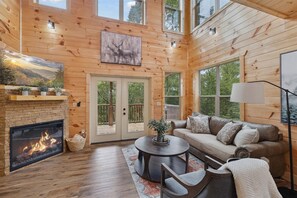 A cozy cabin interior with a fireplace, wooden walls, a sofa, and a large window. - 3 Bedroom with Hot Tub, Games, WiFi, Fire Pit, King Beds + More (Sevierville)