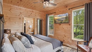 Ein Schlafzimmer einer Holzhütte mit einem Bett, einem Sessel, einem Deckenventilator und großen Fenstern mit Blick auf den Wald.